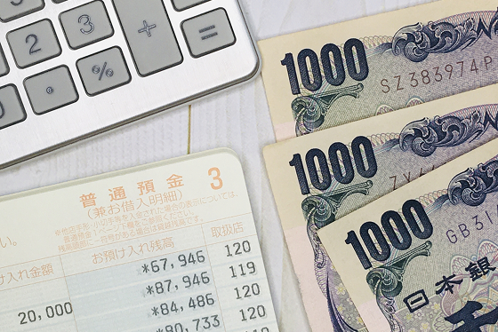 電卓と1000円3枚、銀行の通帳が机に並んでいるイメージの写真。